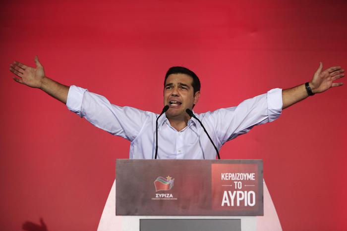 Alexis Tsipras in camicia bianca con le braccia alzate durante una conferenza stampa