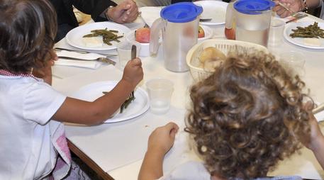 Una mensa scolastica: bambini con posate in mano di fronte ai piatti. Menù