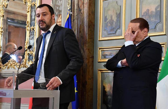 Matteo Salvini sul podio parlando e Silvio Berlusconi dietro con bla mano coprendosi la faccia.