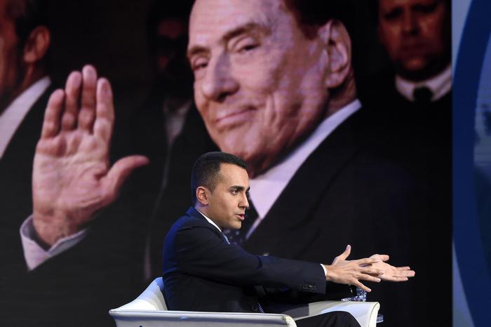 Luigi Di maio seduto in poltrona e sullo sfondo una immagine di Silvio Berlusconi.