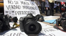 Una macchina fotografica in primo piano e dietro un cartello con la scritta in spagnolo: Non si uccide la verità assassinando i giornalisti". Reporter