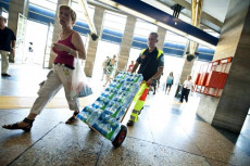 Una signora camminando seguita da un volontario della Protezione Civile con carrettino pieno di bottiglie d'acqua in plastica.