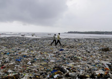 Una spiaggia invasa da bottiglie di plastica e due uomini che passeggiano.