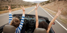 Due ragazze in macchina scoperta su un'autostrada americana alzano le braccia con le dita a V