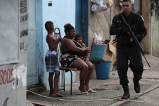 Un soldato armato dei reparti speciali pattuglia una strada. Una donna seduta fuori casa con un bambino.