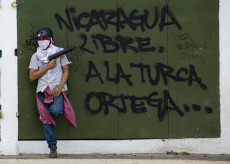 Un ragazzo armato con il viso coperto, appoggiato ad una parete con la scritta "Nicaragua libre. A la turca Ortega...