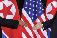 Le mani di Trump e Kim si avvicinano per la storica stretta di mano. Sullo sfondo le bandiere delle due nazioni.