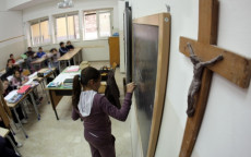 In un'aula di scuola, una bambina scrive sulla lavagna e a lato un crocifisso appeso alla parete.