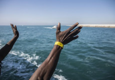 Braccia di migranti tese da una barca in mare, in vista della costa.