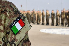 In primo piano la spalla di un militare con la bandiera italiana, sullo sfondo un plotone.