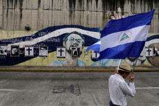 Una persona cammina per strada con la bandiera del Nicaragua e sulla parete sono disegnati i volti di alcuni manifestanti morti