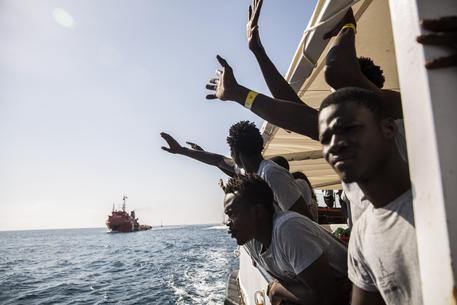 Migranti affacciati sul bordo di una nave salutano sbracciandosi