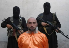 Una persona inginocchiata con la tuta aranciane e alle sue spalle due jidahisti vestiti di nero.