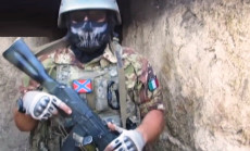 Primo piano di un mercenario in tuta mimetica, con il volto coperto da una maschera, imbracciando il fucile.
