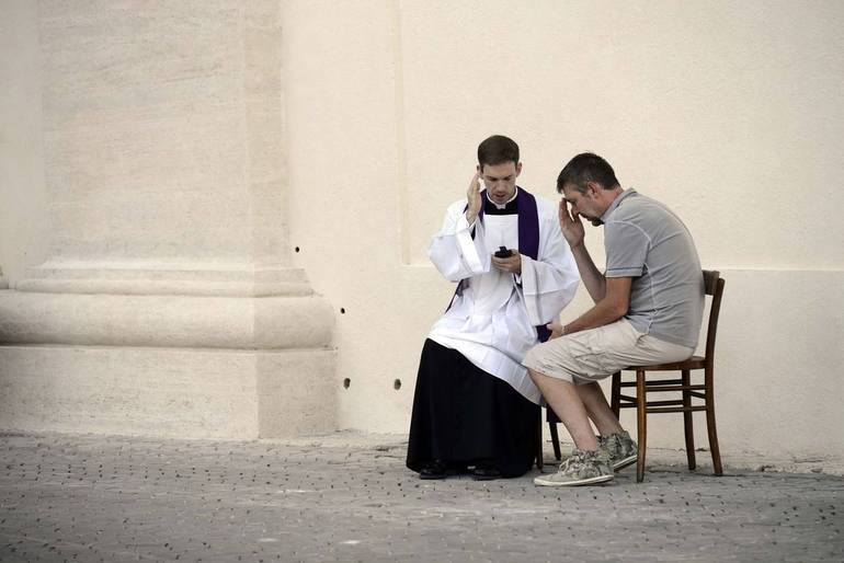 Confessioni all'aperto: un prete ed un fedele seduti su delle sedie nella navata di una chiesa.