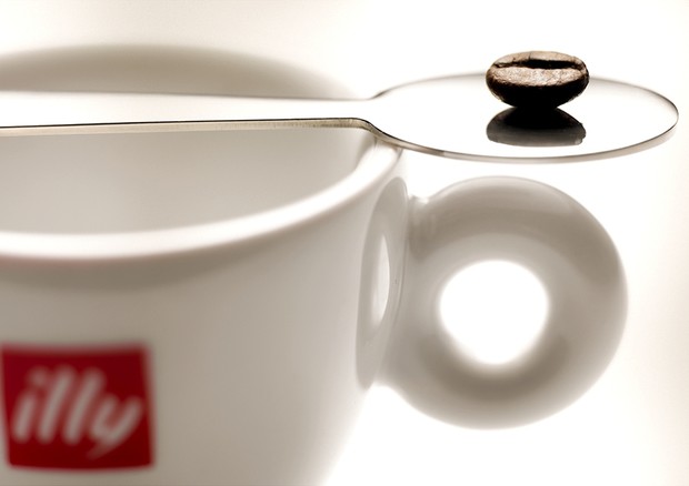 Un chicco di caffè posato su un cucchiaino sopra una tazzina con il logo di Illy