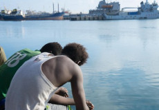 Due migranti seduti sul bordo di una banchina di un porto. Amnesty