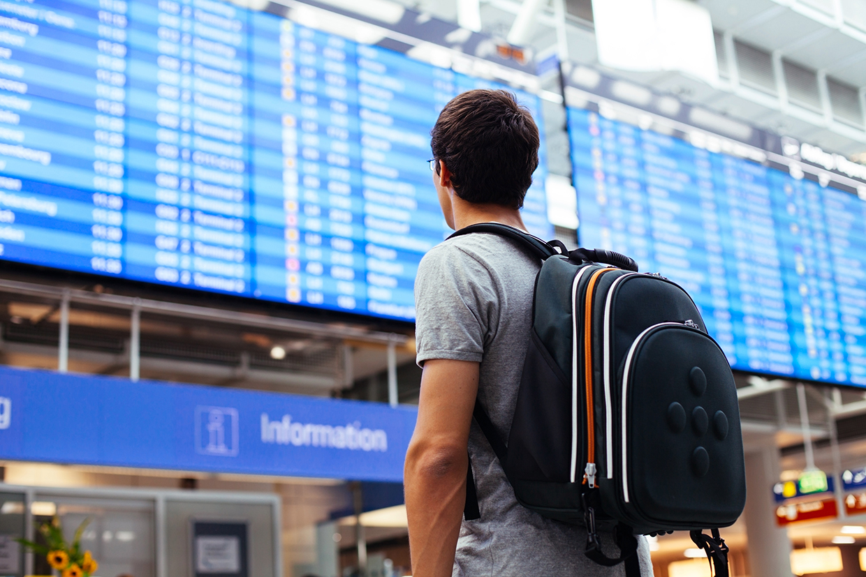 Un ragazzo, zainetto in spalla, guarda il tabellone degli arrivi e partenze in un aeroporto.