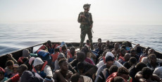 Migranti a bordo di un'imbarcazione controllati da un soldato armato.