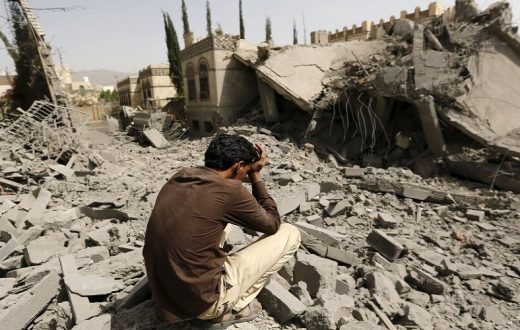 Una pesona seduta a terra con le mani sul viso piange davanti al villaggio distrutto: Bombe italiane in Yemen.