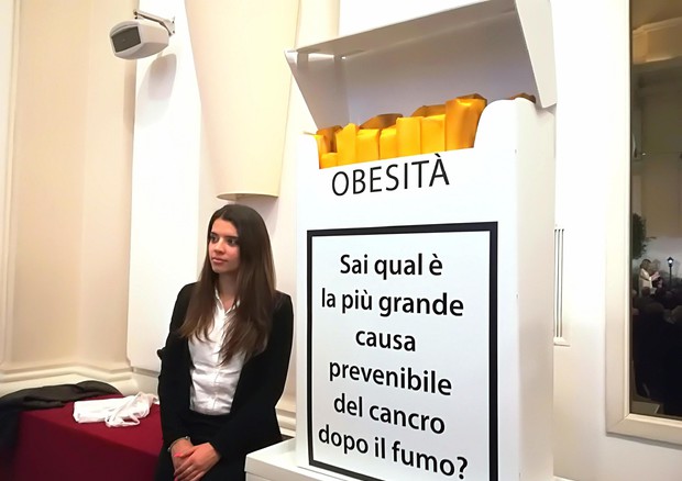 Obesi: Ragazza seduta accanto ad un pacchetto di sigarette gigante con un annuncio del pericolo per la salute dell'obesità.