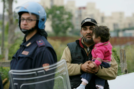Poliziotta controlla lo sgombero di un campo rom. Dietro di lei un padre con bambino in braccio.
