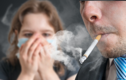 Un uomo con la sigratta in bocca ed una ragazza si copre il viso per non respirare il fumo