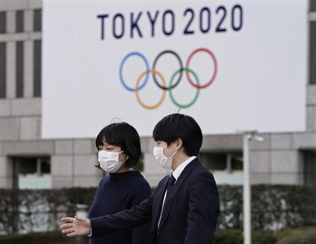 Due passanti con mascherine camminando davanti un cartello in una stazione della metropolitana di Tokyo sulle Olimpiadi Tokyo 2020.