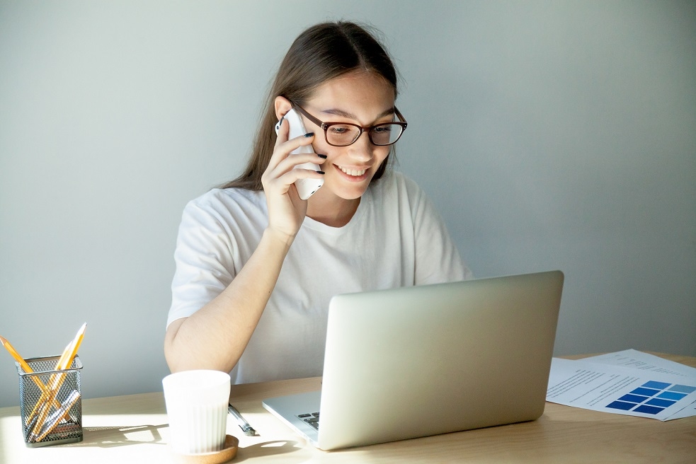 Una giovane donna parla con il telefonino mentre usa la laptop durante il lavoro da casa